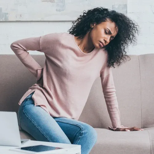 Woman sitting on sofa holding her lower back in visible pain